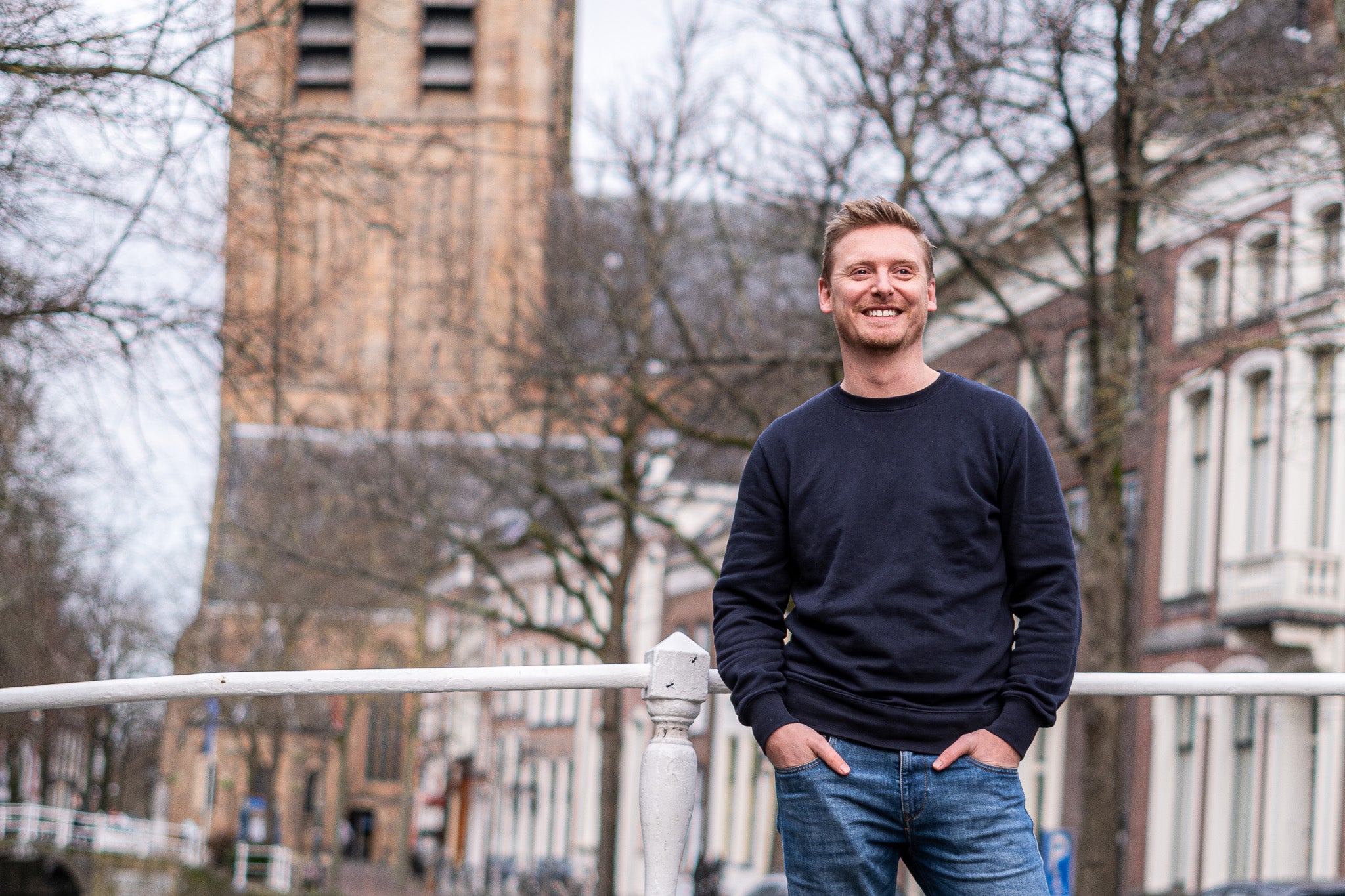 Bas Visser, oprichter van Delft Denim, draagt een donkerblauwe sweater en een spijkerbroek terwijl hij poseert op een brug in Delft, met de historische stad op de achtergrond.