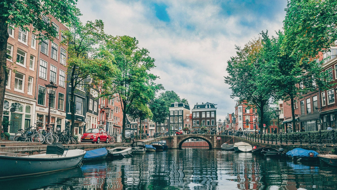 Uitzicht op een pittoreske gracht in Amsterdam, omringd door historische panden en weelderige bomen, met boten die aan de kade liggen en een karakteristieke brug op de achtergrond.