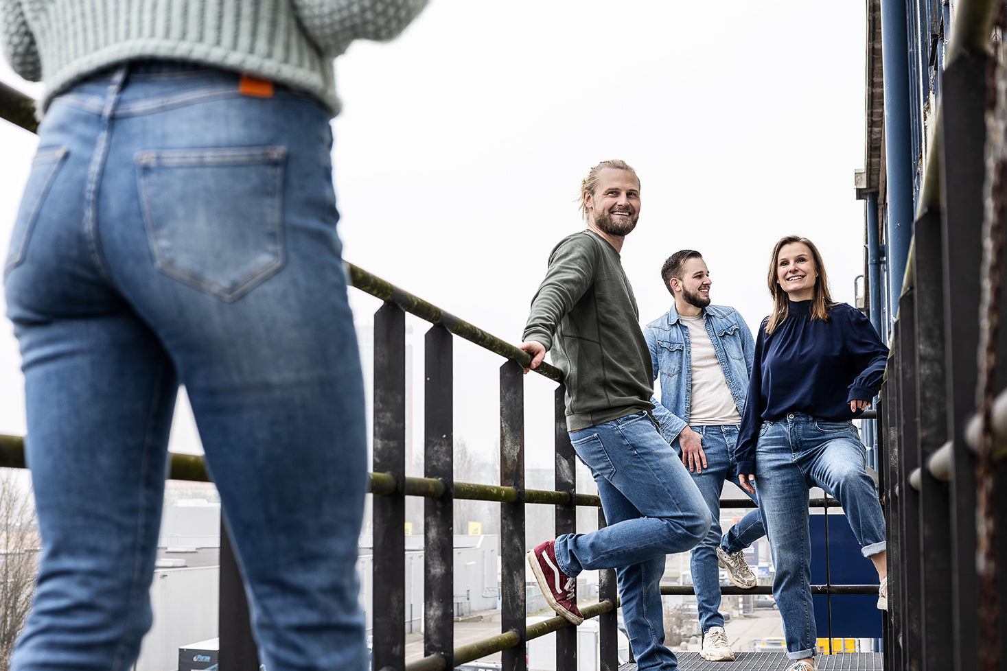 Groep vrienden draagt Delft Denim spijkerbroeken op een industriële locatie. Het voorste model is van achteren te zien, terwijl de andere drie ontspannen poseren en lachen.